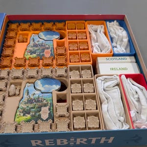 Puede incluir: Caja de juego de mesa abierta, mostrando su contenido. La caja azul tiene la palabra "REBIRTH". Dentro, piezas de juego naranjas, beige y blancas, incluyendo castillos en miniatura. Tarjetas con las etiquetas "SCOTLAND" e "IRELAND".