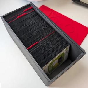May include: A rectangular dark gray storage box filled with a large stack of black trading cards organized by red plastic dividers. A separate red rectangular lid with a cutout design sits beside the open box on a white surface.