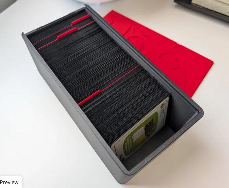 May include: A rectangular dark gray storage box filled with a large stack of black trading cards organized by red plastic dividers. A separate red rectangular lid with a cutout design sits beside the open box on a white surface.