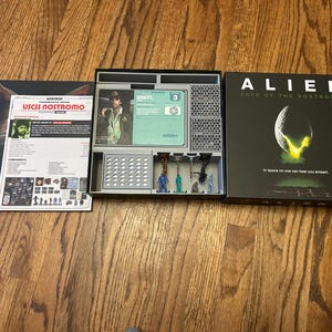 May include: An open board game box set on a wooden surface. The box is black with the title "ALIEN: Fate of the Nostromo" and an image of an alien egg. The set includes game pieces, cards, and a rule book.