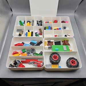 May include: A white plastic organizer with multiple compartments, filled with colorful toy building blocks and accessories. The compartments are arranged in a grid, showcasing various shapes and sizes of blocks, including wheels and other components.