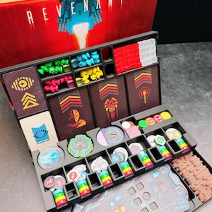 May include: A board game organiser insert for the game Revenant, featuring a black plastic tray with compartments holding various game components including red, green, yellow, and blue tokens, stacks of cards, and small circular markers.