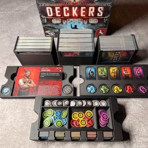 May include: A board game set with the title "DECKERS" on the box. The set includes card holders, cards, tokens, and character cards. The tokens are in various colors, including blue, yellow, and purple. The game appears to be a strategy or role-playing game.