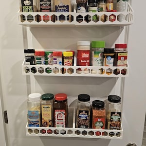 May include: A white, three-tiered spice rack mounted on a door, filled with various spice jars. The jars contain seasonings like garlic, chili, and taco seasoning. The rack has a honeycomb design.