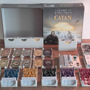 May include: A Game of Thrones Catan board game set. The box is open, revealing game pieces, cards, and a game board. The game pieces are in various colours, including green, blue, yellow, black, purple, and red. The box has the game title and artwork.