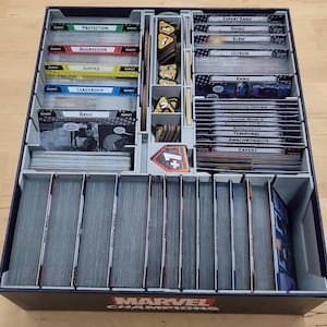 May include: A gray and black plastic organizer with compartments for storing Marvel Champions cards. The organizer has a label that says "Marvel Champions" on the front. The organizer is filled with cards, including "Expert Kang", "Rhino", "Klaw", "Ultron", "Kang", "Legion of Monsters", "Inner Circle", "Temporal Anomalies", "Anachronaunts", "Expert", "Protection", "Aggression", "Justice", "Leadership", and "Basic".