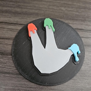 May include: A 3D printed round coaster with a white handprint in the center. The handprint has three raised fingers with drips of red, green, and blue paint.
