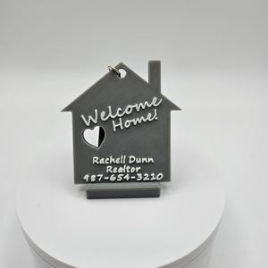 May include: Gray 3D printed house-shaped keychain with the words "Welcome Home!" and a heart cutout. The keychain also includes a business card with the name "Rachell Dunn Realtor" and a phone number.