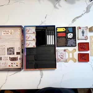 May include: An open board game box with various components laid out. Includes cards, game pieces, dice, and markers. The box lid displays the text "SURVIVE! ALIVE". The game appears to be a strategy or survival-themed board game.