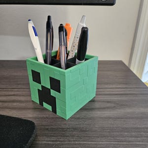 Puede incluir: Un portal&aacute;pices de pl&aacute;stico verde con forma de cara de Creeper de Minecraft. El portal&aacute;pices tiene seis ranuras para bol&iacute;grafos y l&aacute;pices.