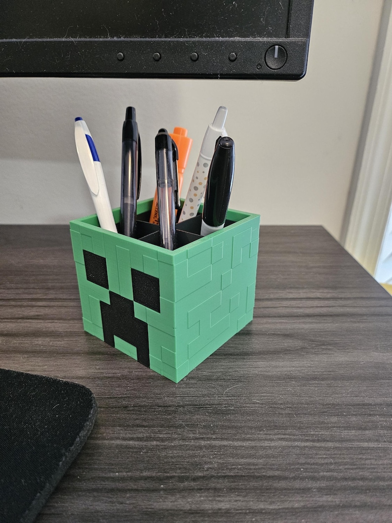 Puede incluir: Un portal&aacute;pices de pl&aacute;stico verde con forma de cara de Creeper de Minecraft. El portal&aacute;pices tiene seis ranuras para bol&iacute;grafos y l&aacute;pices.