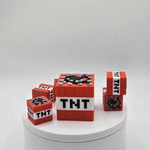 May include: Five red and black Minecraft TNT blocks. The largest block has the word "TNT" printed on it. The smaller blocks have the word "TNT" printed on the side.