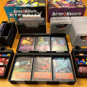 May include: A set of black and gray plastic organizers for a board game. The organizers are filled with cards, dice, and tokens. The game is called Astro Knights: Eternity.
