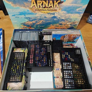 Puede incluir: Caja de juego de mesa abierta, con el título "Arnak" en la tapa. En el interior, organizadores de plástico negro contienen cartas, fichas y piezas del juego. Un mapa y una brújula son visibles en el lateral.