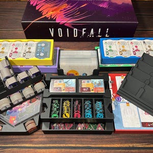 May include: A board game titled "VOIDFALL" with various game components. The components include colorful trays, cards, and tokens. The game box is purple with the game title in white. The game components are arranged on a wooden surface.