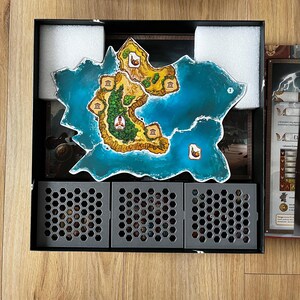 Puede incluir: Un tablero de juego con un mapa azul y verde de una isla con varios edificios. El tablero está rodeado de espuma gris y tres compartimentos de almacenamiento hexagonales grises.