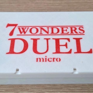 Può includere: Una scatola rettangolare bianca con la scritta "7 WONDERS DUEL micro" in rosso. La scatola ha un coperchio leggermente rialzato e piccole rientranze sul bordo anteriore. La scatola del gioco è su una superficie marrone chiaro.