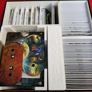 May include: A white plastic organizer with compartments for storing cards and tokens. The organizer has labels for different card types, including "Barracks - Treasures", "Level 3 Treasures", "Level 2 Treasures", "Level 1 Treasures", "Serene", "Phadraxa", "Ohat and Blumor", "Mist: Aeon's End", and "Indira".