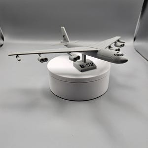 May include: A gray B-52 model airplane, displayed on a white circular stand. The model has detailed wings, engines, and a tail fin. The base of the model reads "B-52". The airplane is a detailed replica of the iconic bomber.
