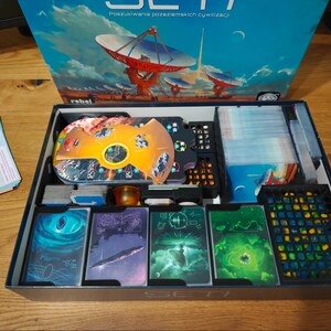 May include: A board game box set with the title "SETI" and images of a satellite dish and planets. The open box reveals game components, including cards with colorful illustrations, a game board, and other game pieces. The box is on a wooden surface.