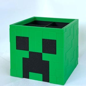 Puede incluir: Una caja de almacenamiento de pl&aacute;stico verde con forma de cara de Creeper de Minecraft. La caja tiene cuatro compartimentos y es perfecta para organizar objetos peque&ntilde;os.
