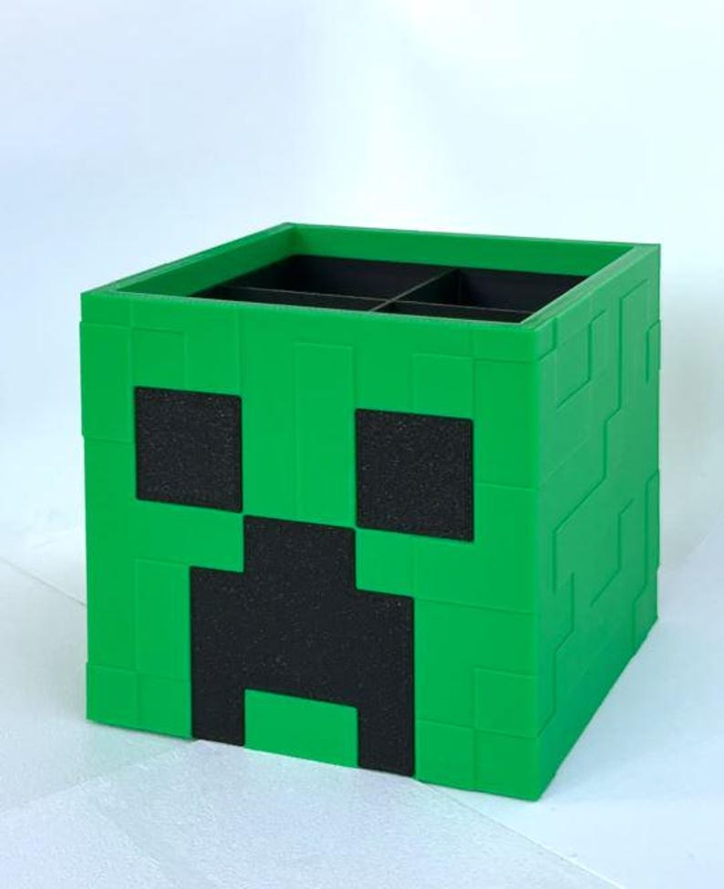 Puede incluir: Una caja de almacenamiento de pl&aacute;stico verde con forma de cara de Creeper de Minecraft. La caja tiene cuatro compartimentos y es perfecta para organizar objetos peque&ntilde;os.