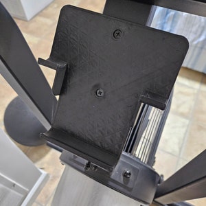 May include: Black plastic phone holder attached to a black exercise machine. The holder has two screws on the back and a small lip on the side.