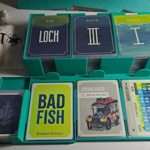 May include: A teal card game organiser filled with various game cards. The cards feature illustrations and text, including "The Loch," "Depth III," and "Bad Fish." A small canvas bag with "CLOUT" printed on it is also visible.