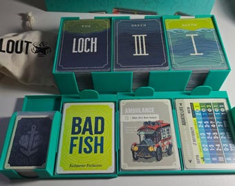 May include: A teal card game organizer filled with various game cards. The cards feature illustrations and text, including "The Loch," "Depth III," and "Bad Fish." A small canvas bag with "CLOUT" printed on it is also visible.