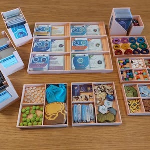 May include: A collection of pink game organizers filled with game components. The organizers hold cards, tokens, and other game pieces. The components are in various colors and shapes, including blue, green, yellow, and brown.