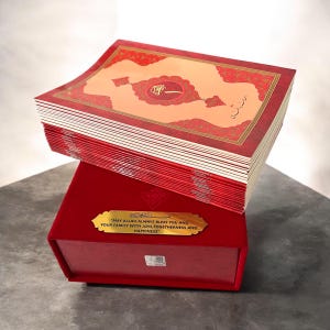 Könnte beinhalten: Ein rotes und goldenes Buch mit kunstvollen Designs, das auf einer passenden roten Box liegt. Die Seiten des Buches sind sichtbar, und die Box hat eine goldene Plakette mit Text. Der Text auf der Plakette lautet: "MAY ALLAH ALWAYS BLESS YOU AND YOUR FAMILY WITH JOYS, TOGETHERNESS AND HAPPINESS."