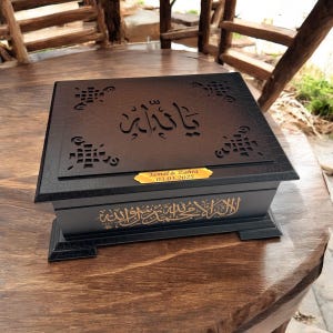 Puede incluir: Caja de madera negra con intrincados diseños calados en la tapa y la base. La caja presenta letras doradas y una pequeña placa dorada con los nombres "Jamal & Zahra" y la fecha "03.03.2025".
