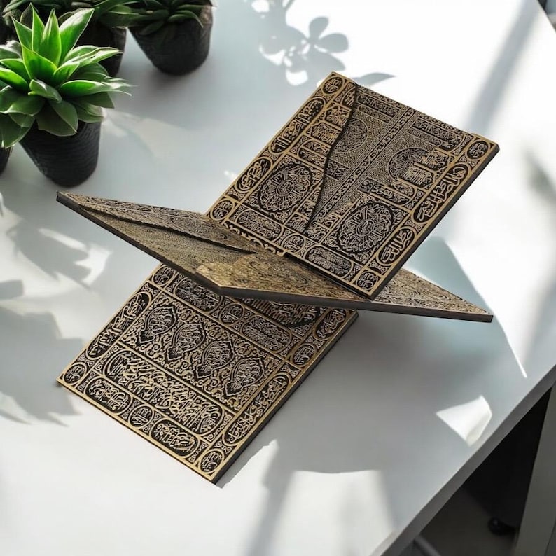 May include: A wooden book stand with intricate black and gold patterns. The stand is designed to hold a book open, with a three-panel design. The stand is on a white surface, with small potted plants in the background.