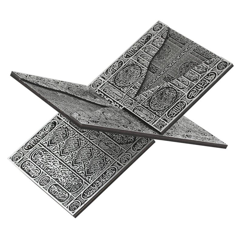 May include: A silver-colored book stand with intricate, engraved patterns and Arabic calligraphy. The stand is designed to hold a book open, with multiple panels angled for display. The design is detailed and ornate.