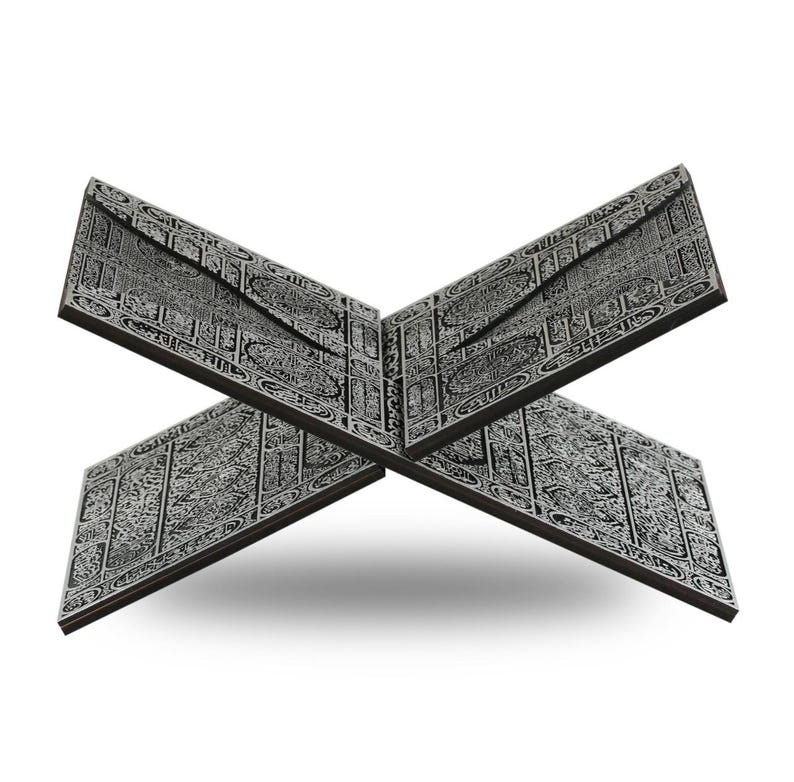 May include: A dark gray wooden book stand with intricate, engraved patterns. The stand is designed to hold a book open, with two panels forming an X shape. The detailed carvings are in a lighter gray, creating a contrast against the darker background.