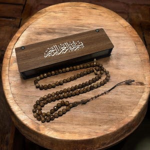 Könnte beinhalten: Ein hölzernes Gebetsperlenset mit passender Box. Die rechteckige Box hat eine weiße arabische Kalligraphie auf dem Deckel. Die hellbraunen Perlen sind auf einer Holzoberfläche angeordnet.
