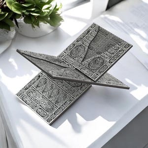 May include: A silver-colored book stand with intricate, engraved patterns. The stand is designed to hold a book open, with three angled panels. The design features detailed calligraphy and geometric shapes, creating an elegant and decorative piece.