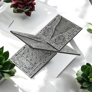 May include: A silver-colored, open book with intricate, engraved patterns and Arabic script. The book is displayed on a white surface, surrounded by green and red plants. The pages are slightly angled, creating a dynamic composition.