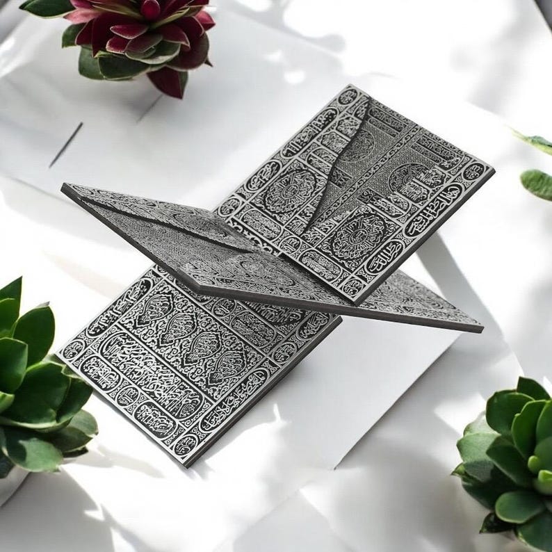 May include: A silver-colored, open book with intricate, engraved patterns and Arabic script. The book is displayed on a white surface, surrounded by green and red plants. The pages are slightly angled, creating a dynamic composition.