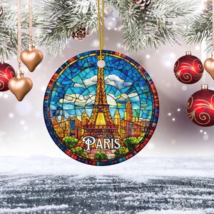 May include: Round Christmas ornament featuring a stained-glass design of the Eiffel Tower and the city of Paris. The ornament has a gold string for hanging. The word "PARIS" is written across the bottom. Red and gold ornaments hang in the background.