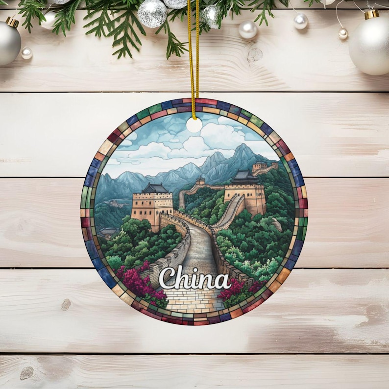 May include: Round Christmas ornament featuring a stained-glass style illustration of the Great Wall of China. The image shows a section of the wall winding through green mountains under a partly cloudy sky.  The colors are predominantly greens, blues, and earth tones, with a multicolored border. The word 'China' is written in a stylized script at the bottom of the ornament.