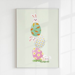 May include: Framed print showing three decorated Easter eggs stacked with cartoon bunnies. The top egg is green with orange carrot designs, the middle egg has yellow polka dots, and the bottom egg is pink with white flowers.