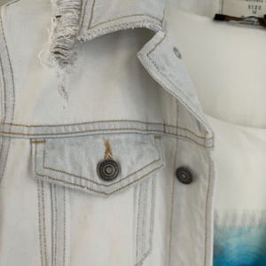 Puede incluir: Una chaqueta vaquera azul claro con un detalle desgastado en el cuello y un solo bolsillo en el lado izquierdo. La chaqueta tiene cierre de botones y una etiqueta que dice "Colin's Jeans" y "Talla M".