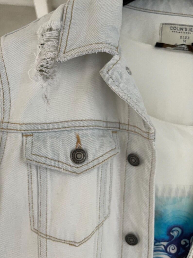 Puede incluir: Una chaqueta vaquera azul claro con un detalle desgastado en el cuello y un solo bolsillo en el lado izquierdo. La chaqueta tiene cierre de botones y una etiqueta que dice "Colin's Jeans" y "Talla M".