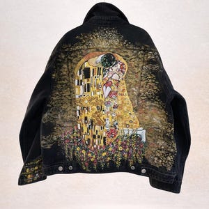 Könnte beinhalten: Schwarze Jeansjacke mit einem Gemälde von Gustav Klimts "Der Kuss" auf dem Rücken. Das Kunstwerk enthält Gold-, Gelb-, Rot- und Blumenelemente. Die Jacke hat einen klassischen Kragen und Knopfverschlüsse. Der Text "DESIGNS DENIM" ist unten aufgedruckt.