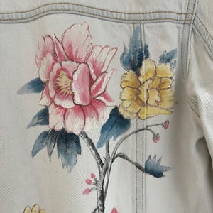 Puede incluir: Una chaqueta vaquera azul claro con un dise&ntilde;o floral pintado a mano. Las flores son rosas, amarillas y azules, y las ramas son negras.
