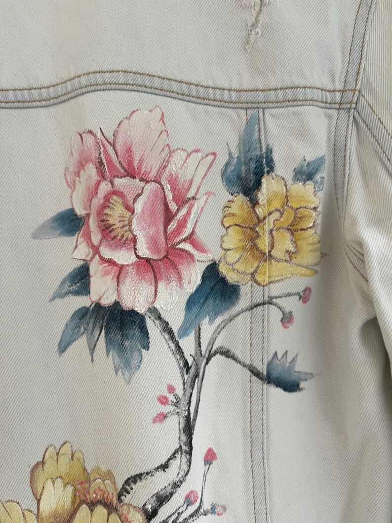 Puede incluir: Una chaqueta vaquera azul claro con un dise&ntilde;o floral pintado a mano. Las flores son rosas, amarillas y azules, y las ramas son negras.