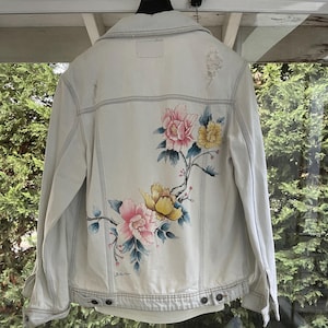 Puede incluir: Una chaqueta vaquera azul claro con un dise&ntilde;o floral pintado en la espalda. Las flores son rosas, amarillas y blancas. La chaqueta tiene un aspecto desgastado con algunos desgarros y roturas.
