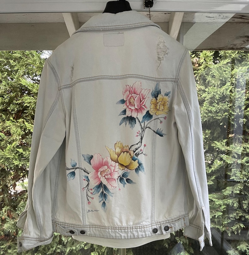 Puede incluir: Una chaqueta vaquera azul claro con un dise&ntilde;o floral pintado en la espalda. Las flores son rosas, amarillas y blancas. La chaqueta tiene un aspecto desgastado con algunos desgarros y roturas.
