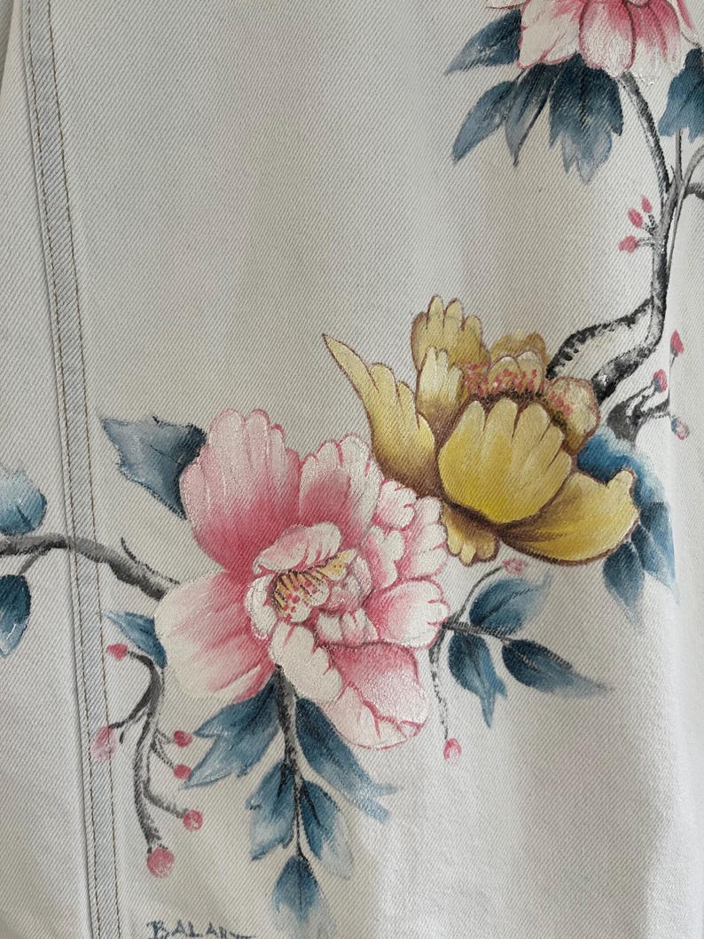 Puede incluir: Una chaqueta vaquera blanca con dise&ntilde;os florales pintados a mano. Las flores son rosas, amarillas y azules. La chaqueta tiene una textura sutil.